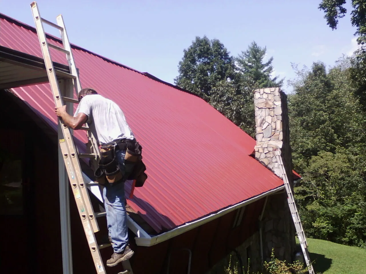 Roofer on a ladder beside a red metal roof and stone chimney — Soffit & Fascia Repair by Roofing Brothers in West Friendship